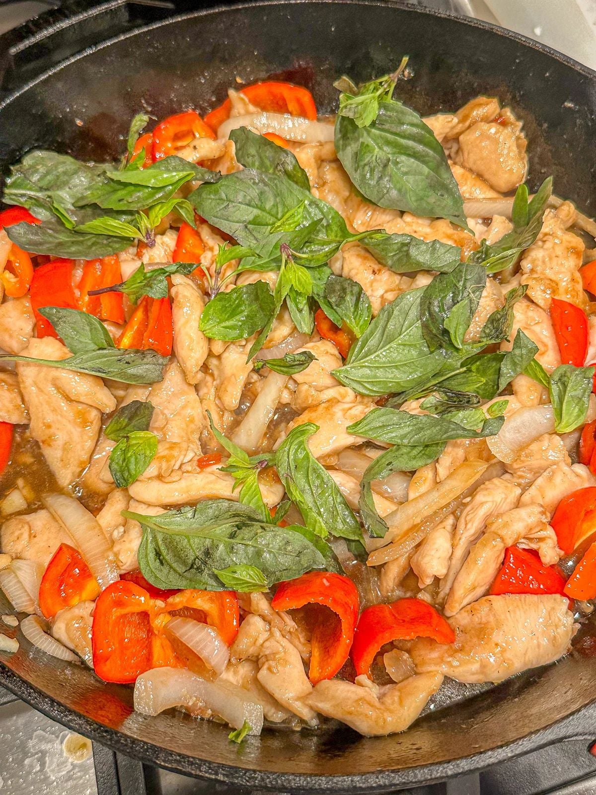 Stir-fried chicken, red bell peppers, and onions topped with fresh basil leaves in a hot pan.