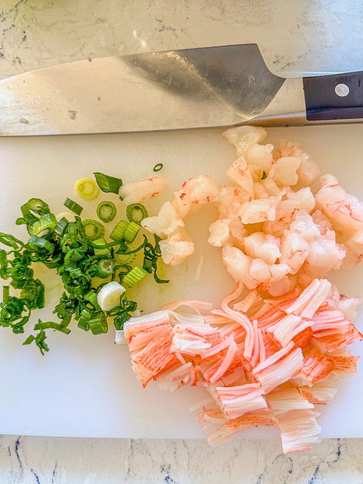 Sliced green onions, shrimp, and imitation crab arranged on a cutting board with a knife placed beside them.
