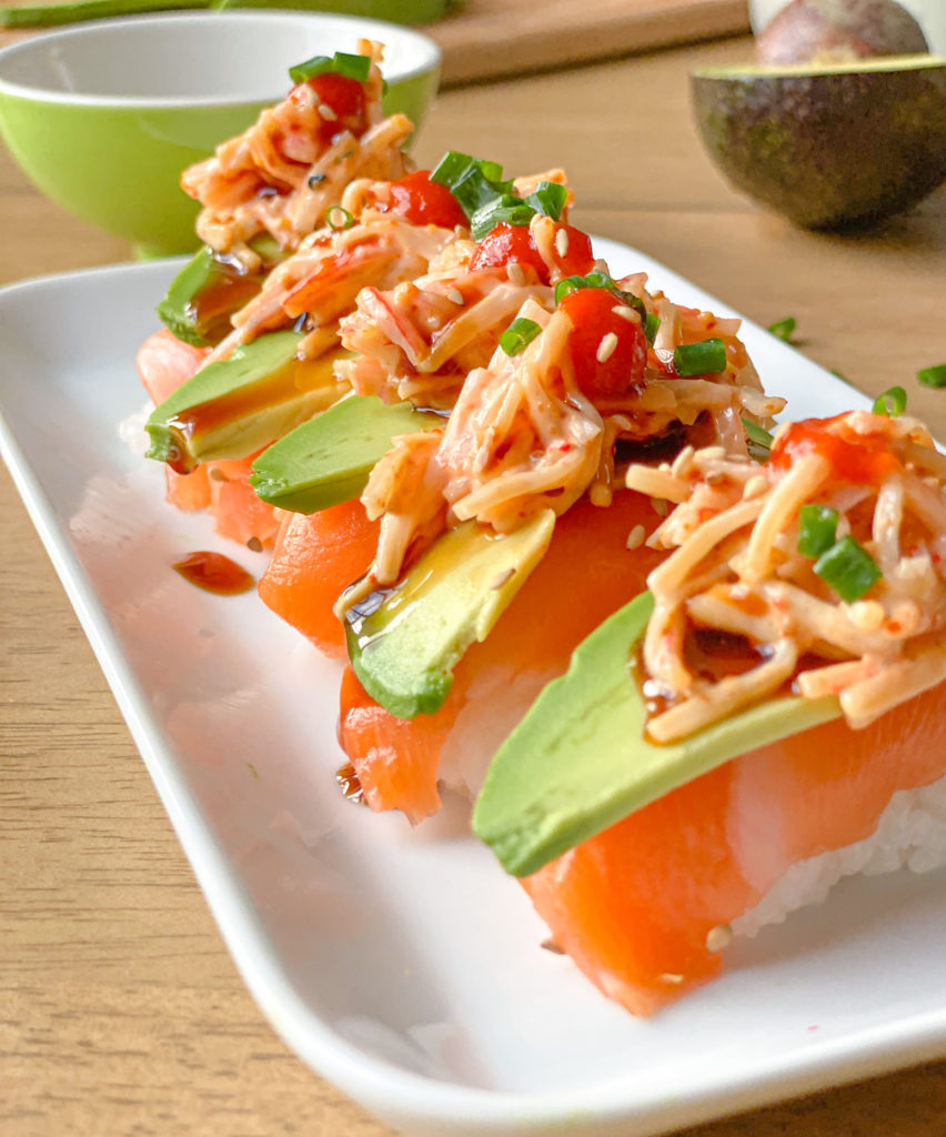 salmon thin slices on top of sushi rice, thin avocado slices, imitation crab spicy mayo and green onions on top