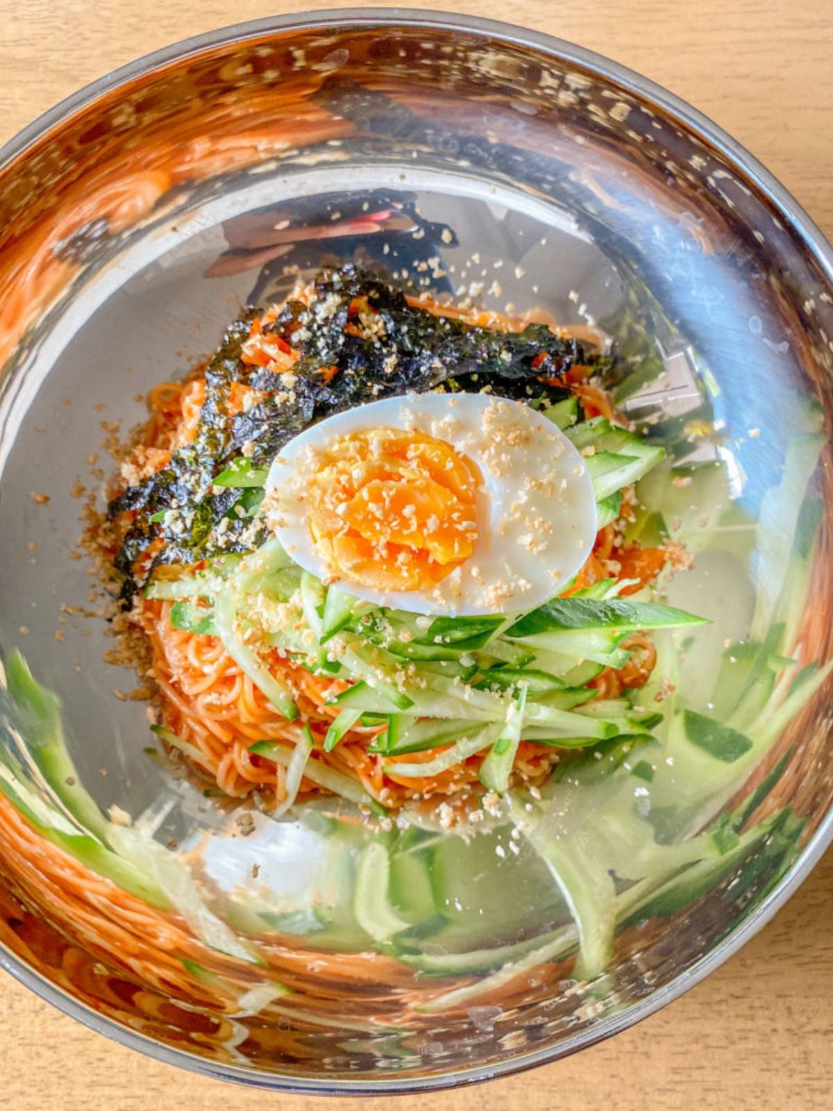 Spicy Korean cold noodles in a bowl topped with an egg and sliced cucumbers.