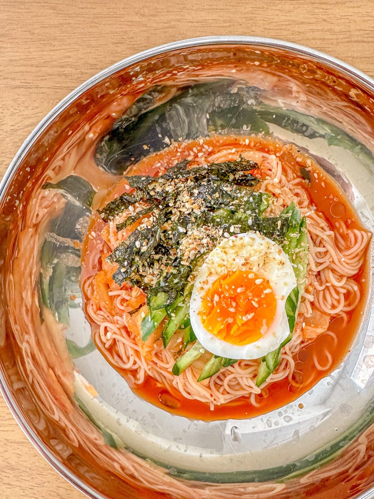 Spicy Korean cold noodles in a bowl topped with an egg and sliced cucumbers.