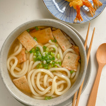 Udon soup with fish cakes in a bowl topped with green onions and tempura shrimp on the side.