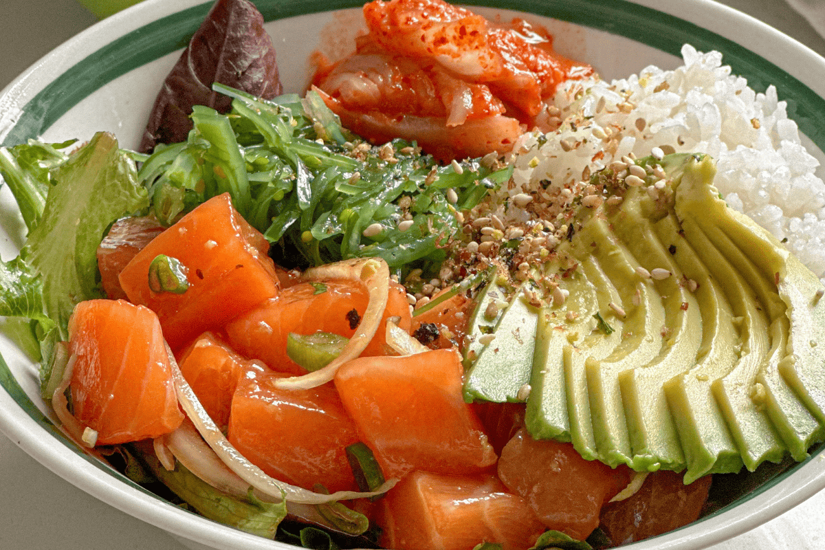 avocado sliced with salmon poke, white rice, seaweed salad, salad, kimchi in a bowl served with soy sauce and chopsticks