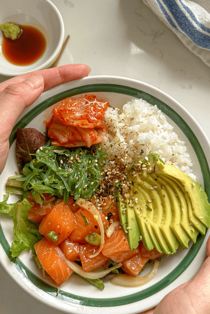 salmon poke, sliced avocado, seasweed salad, lettuce and white rice in a bowl with furikake seasoning on top with hands holding the side