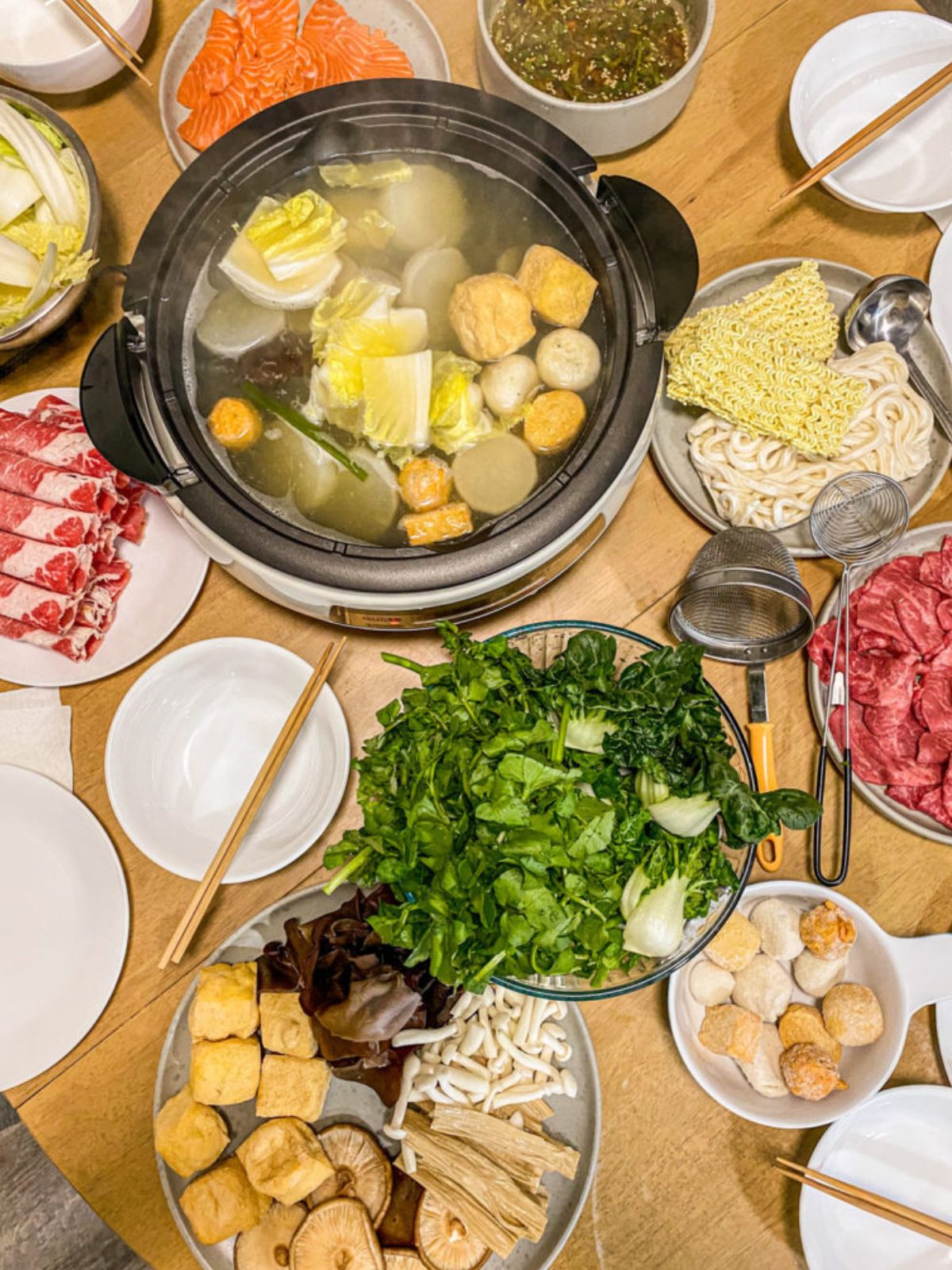 Hot pot on dining table filled with vegetables, ramen, tofu, mushrooms, raw beef, and fish balls.