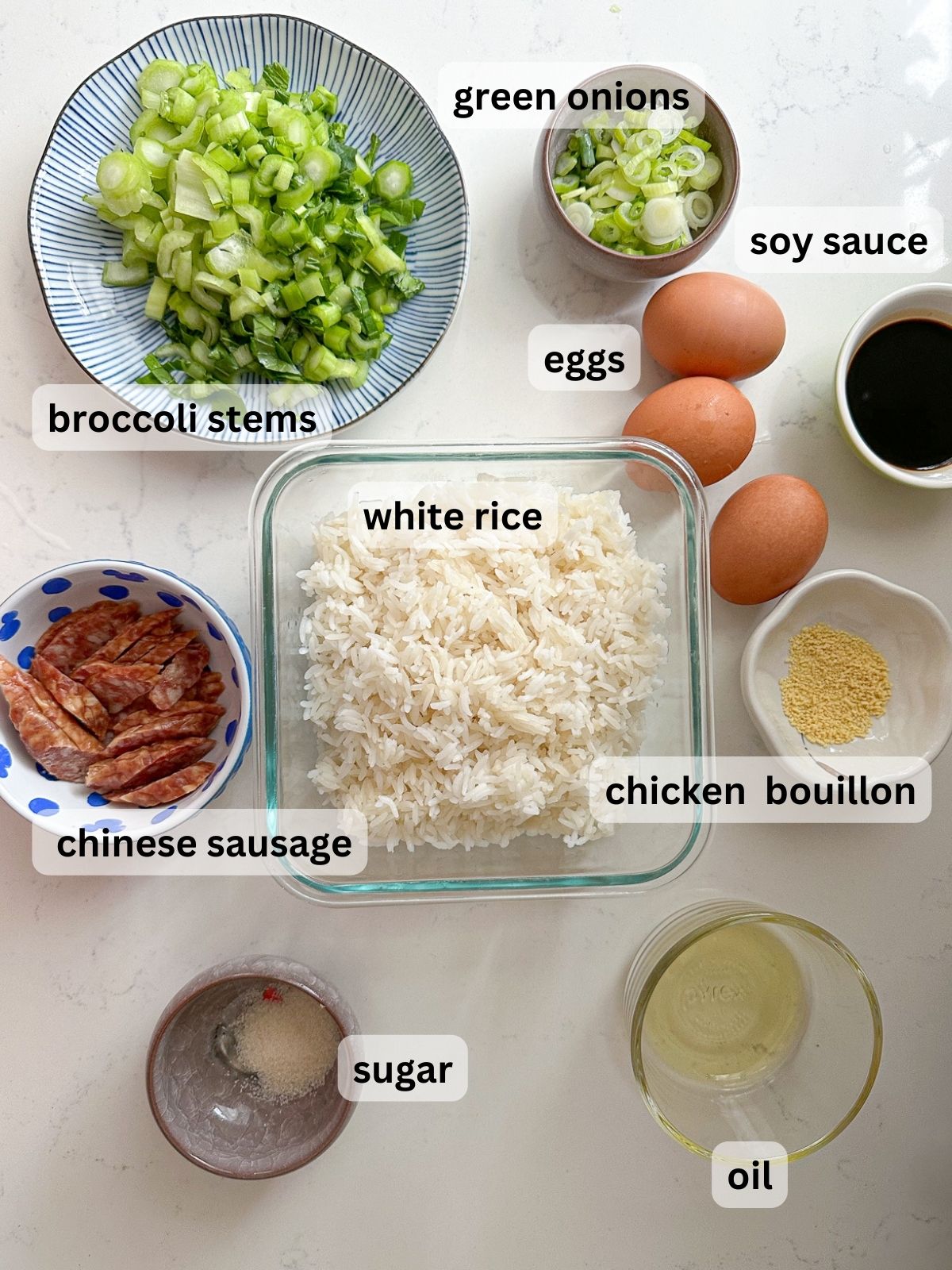 Ingredients to make golden fried rice include broccoli stems, white rice, eggs, green onions, soy sauce, chinese sausage chicken bouillon, oil, and sugar on the table with text labels.