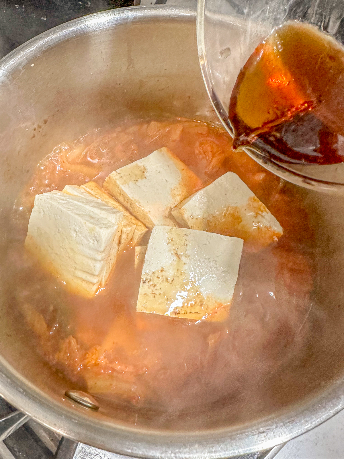 Adding soy sauce to a pot of boiling tofu, kimchi and broth.