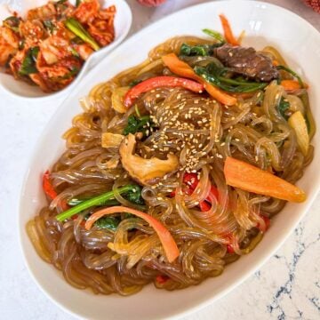 A plate of glass noodles, red bell peppers, spinach, and sprinkled sesame seeds, with a small dish of kimchi.