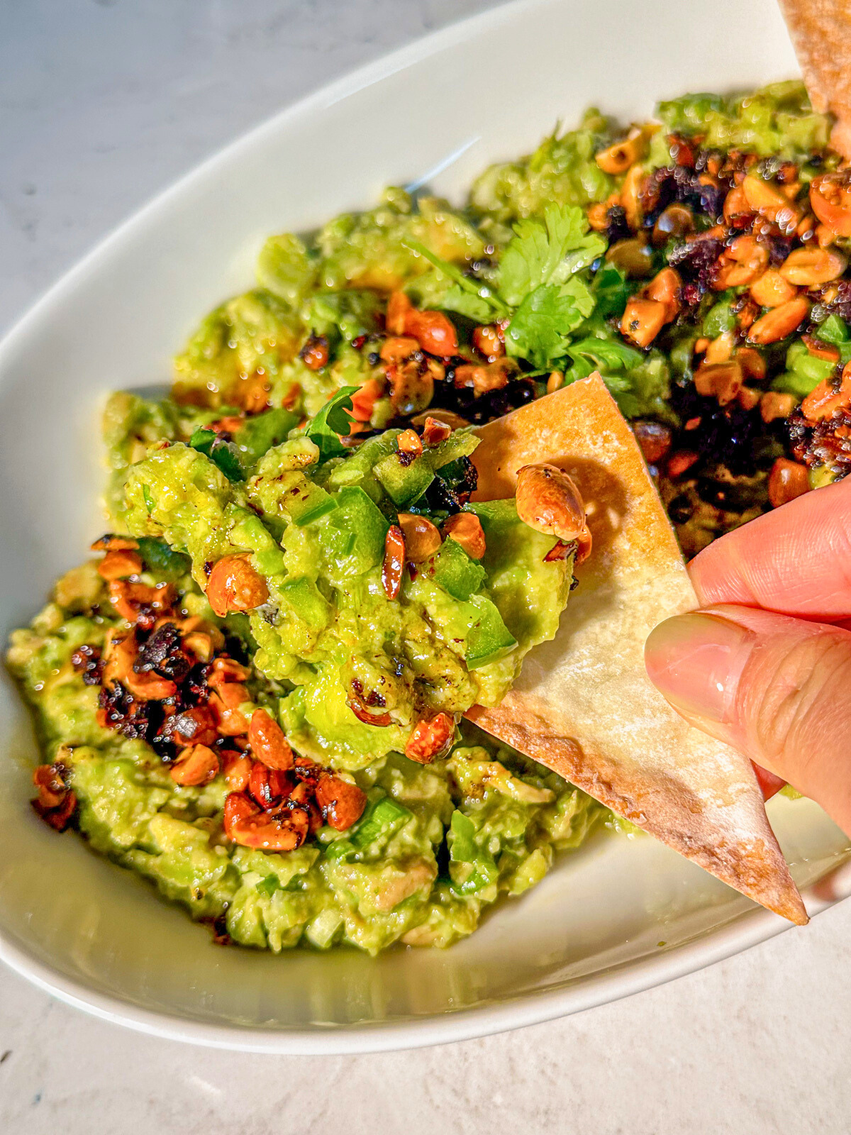 A bowl of guacamole with sprinkled garlic and nuts , along with a taco piece on hand with guac on top.