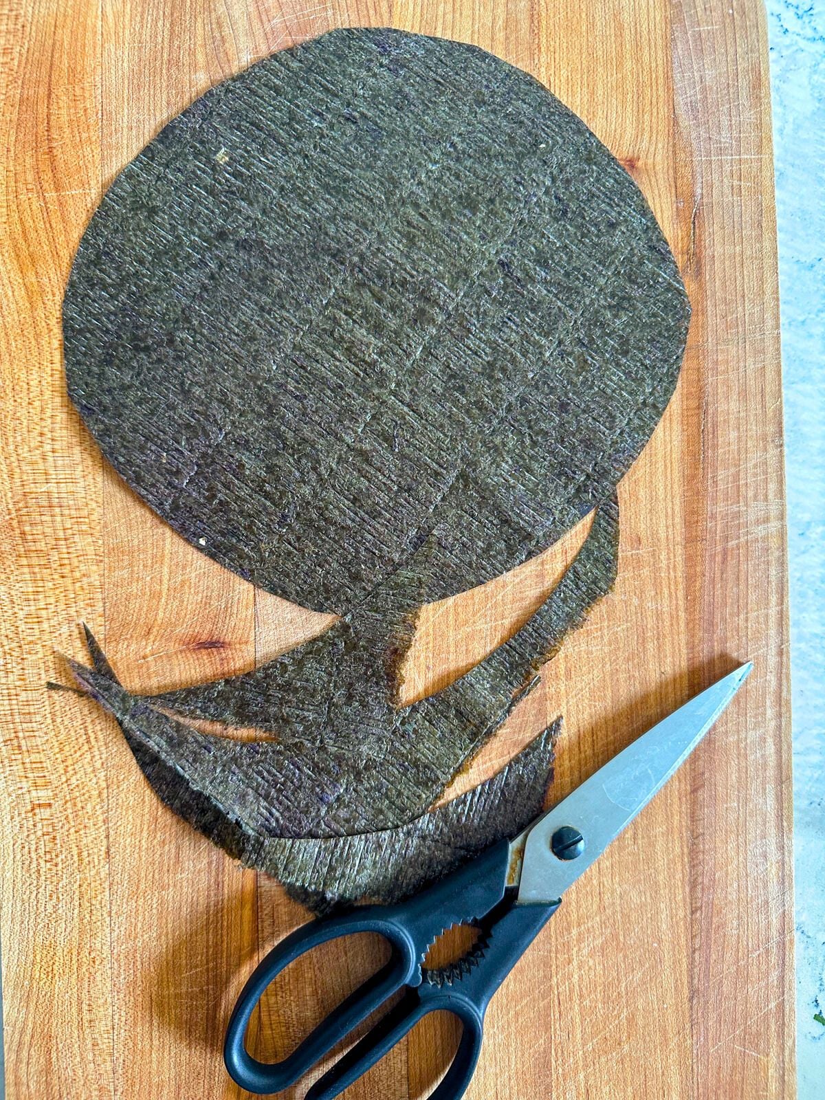 A piece of circle shaped seaweed (nori) on a chopping board next to a kitchen knife.