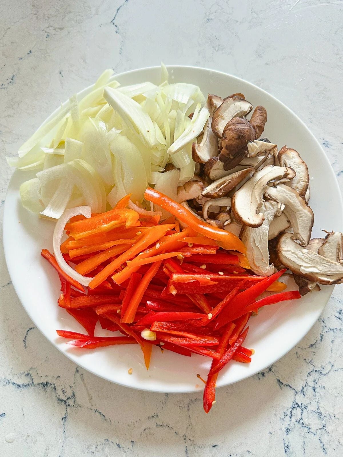 A plate of sliced vegetables including red bell peppers, yellow onion, and shiitake mushrooms, prepared for cooking.
