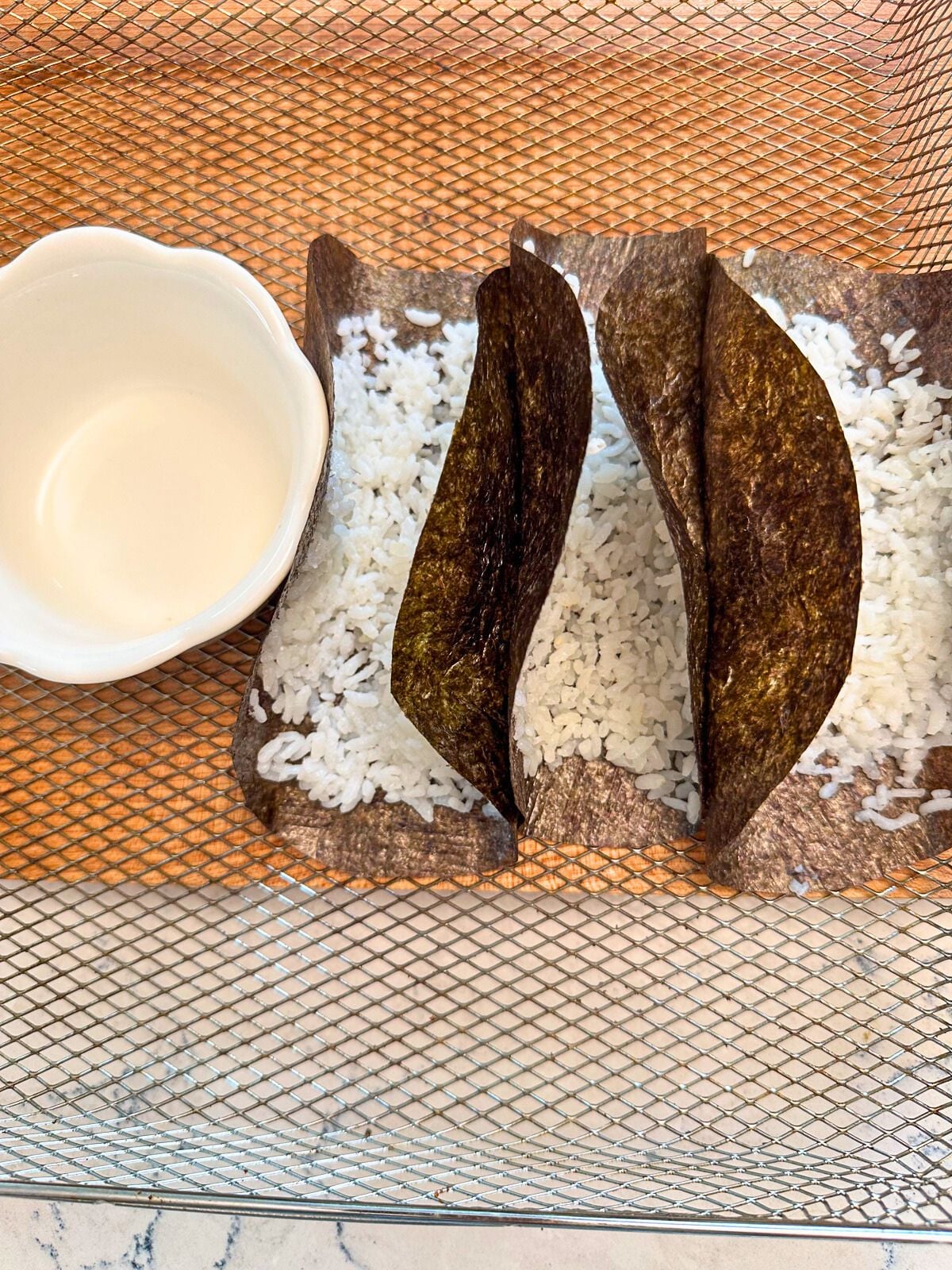 Three folded seaweeds with cooked white rice positioned next to a tiny bowl on a metal tray.
