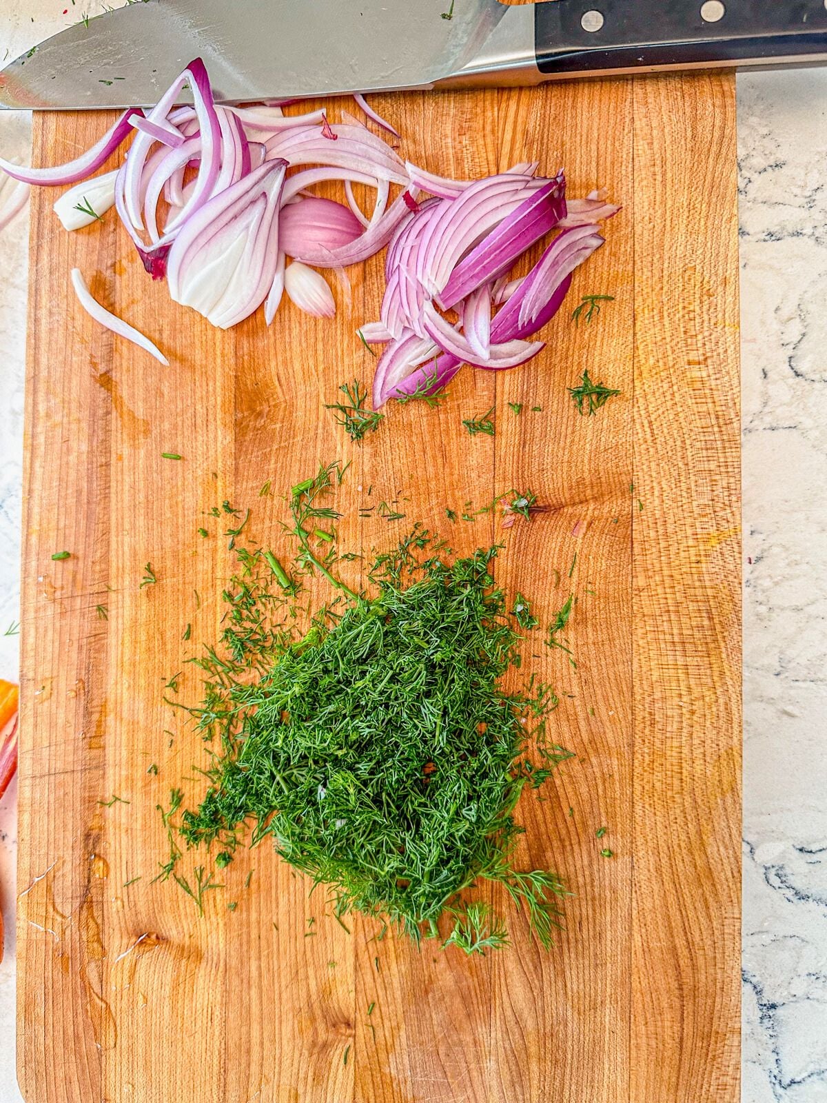 Thin slices of red onion and chopped dill leaves on a chopping board with a kitchen knife on the side.