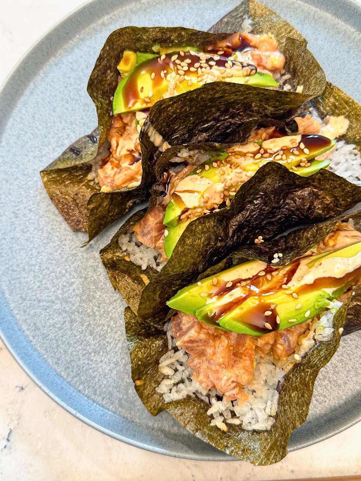 Three Tuna Poke Tacos on a plate with teriyaki sauce, avocado slices, and sesame seeds on top.