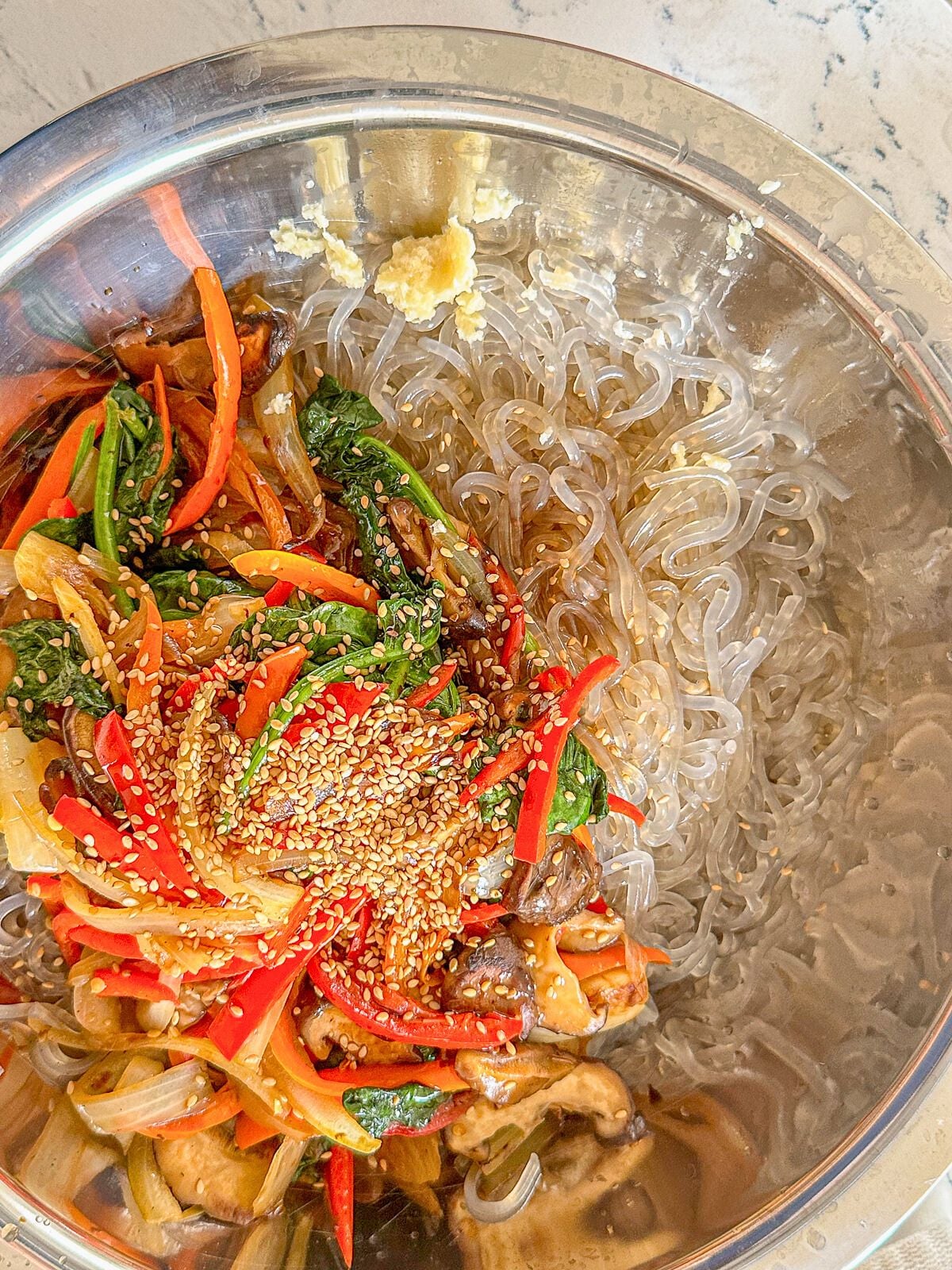 Korean glass noodles in a large metal bowl topped with stir-fried vegetables, garlic, sauce, and sprinkled with sesame seeds.