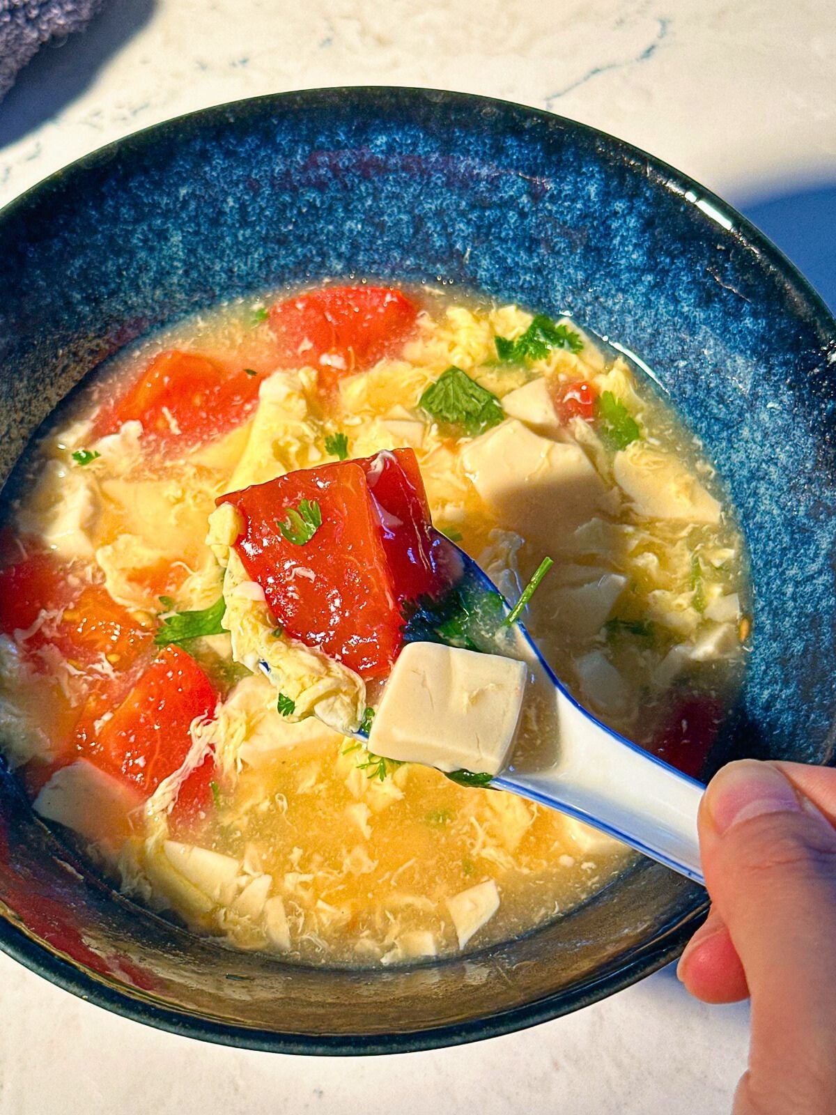 Scooping some tomato egg soup with tofu chunks and sprinkled cilantro from a bowl.