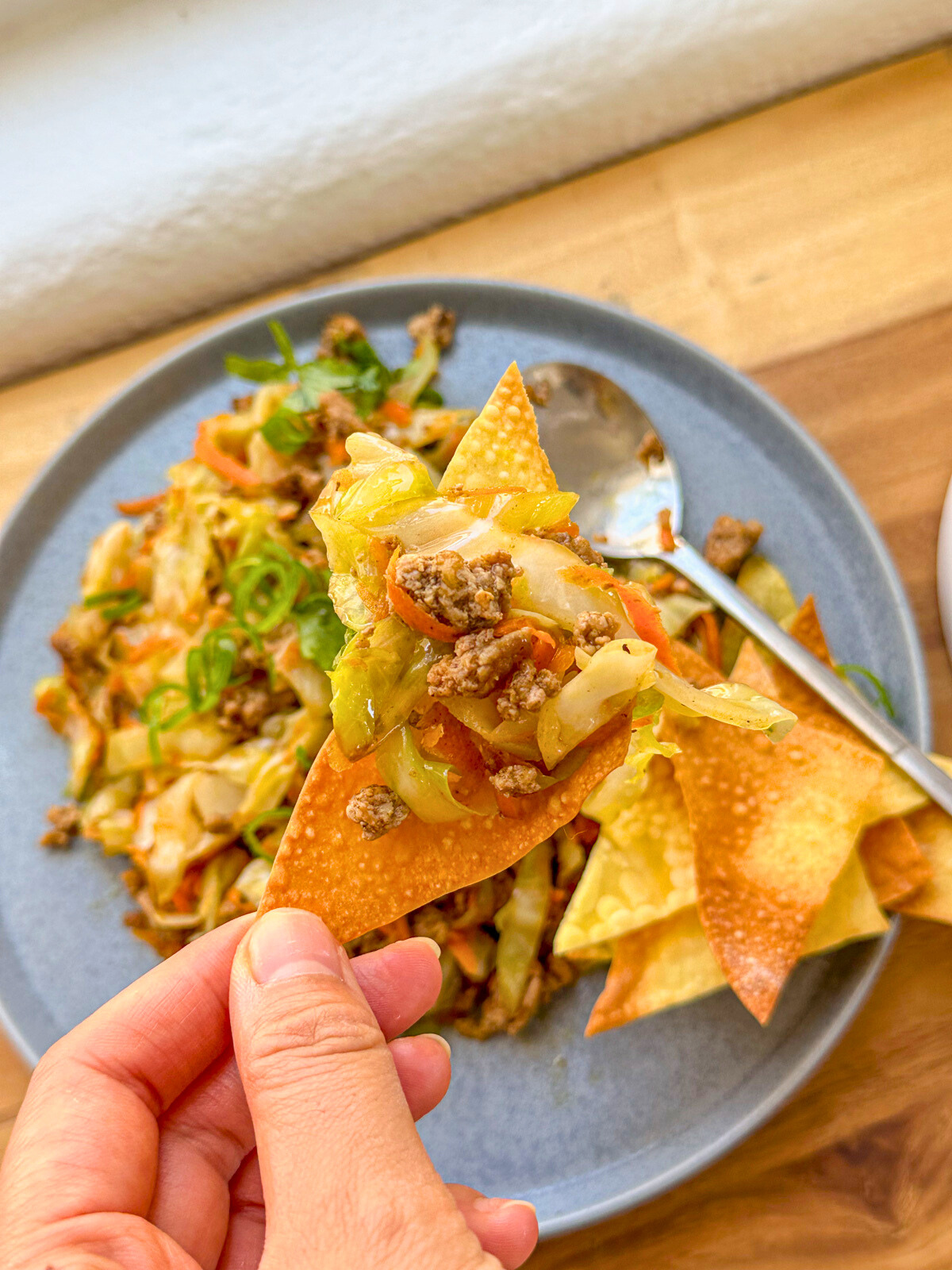Holding a wonton chip topped with egg roll filling, with a plate of garnished egg roll in a plate in the background.