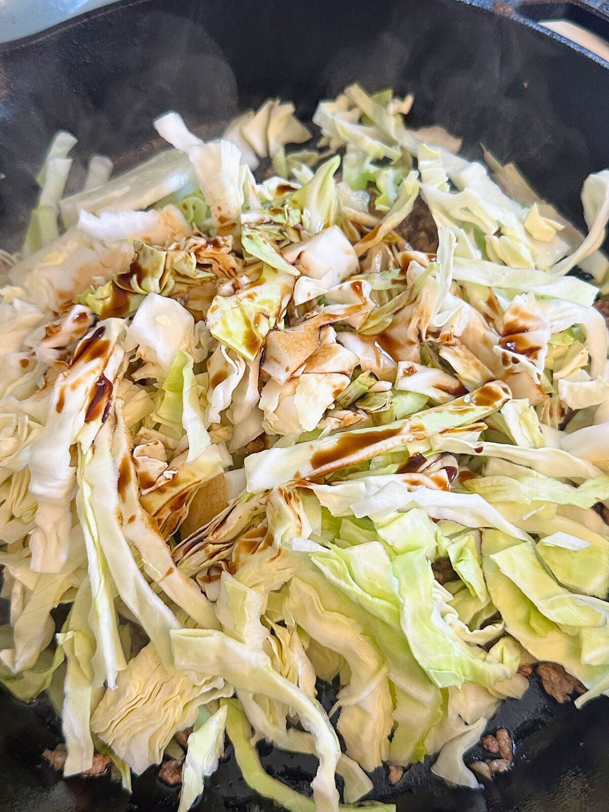 Cabbage and soy sauce being added to a pan with cooked ground pork.