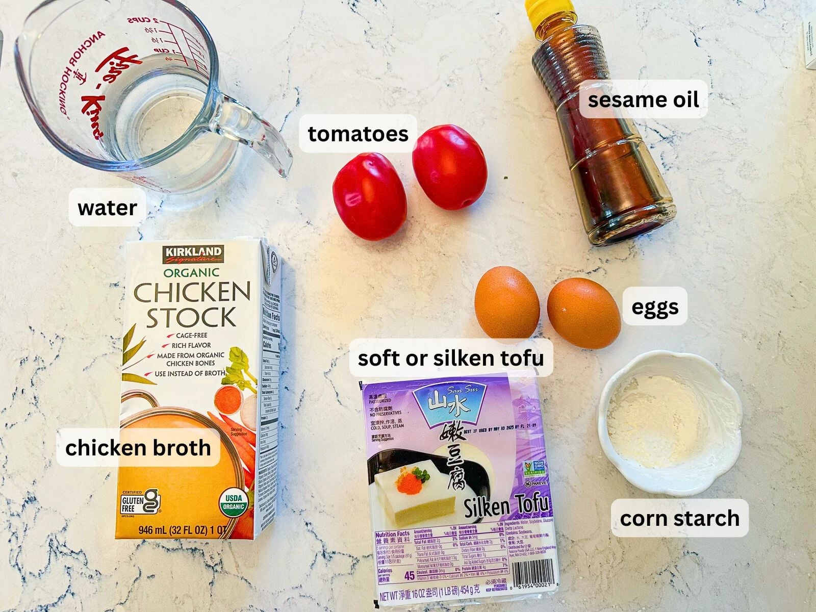 List of tomato egg soup ingredients including water, tomatoes, eggs, sesame oil, boxed chicken broth, a small dish of cornstarch, and packaged soft tofu.