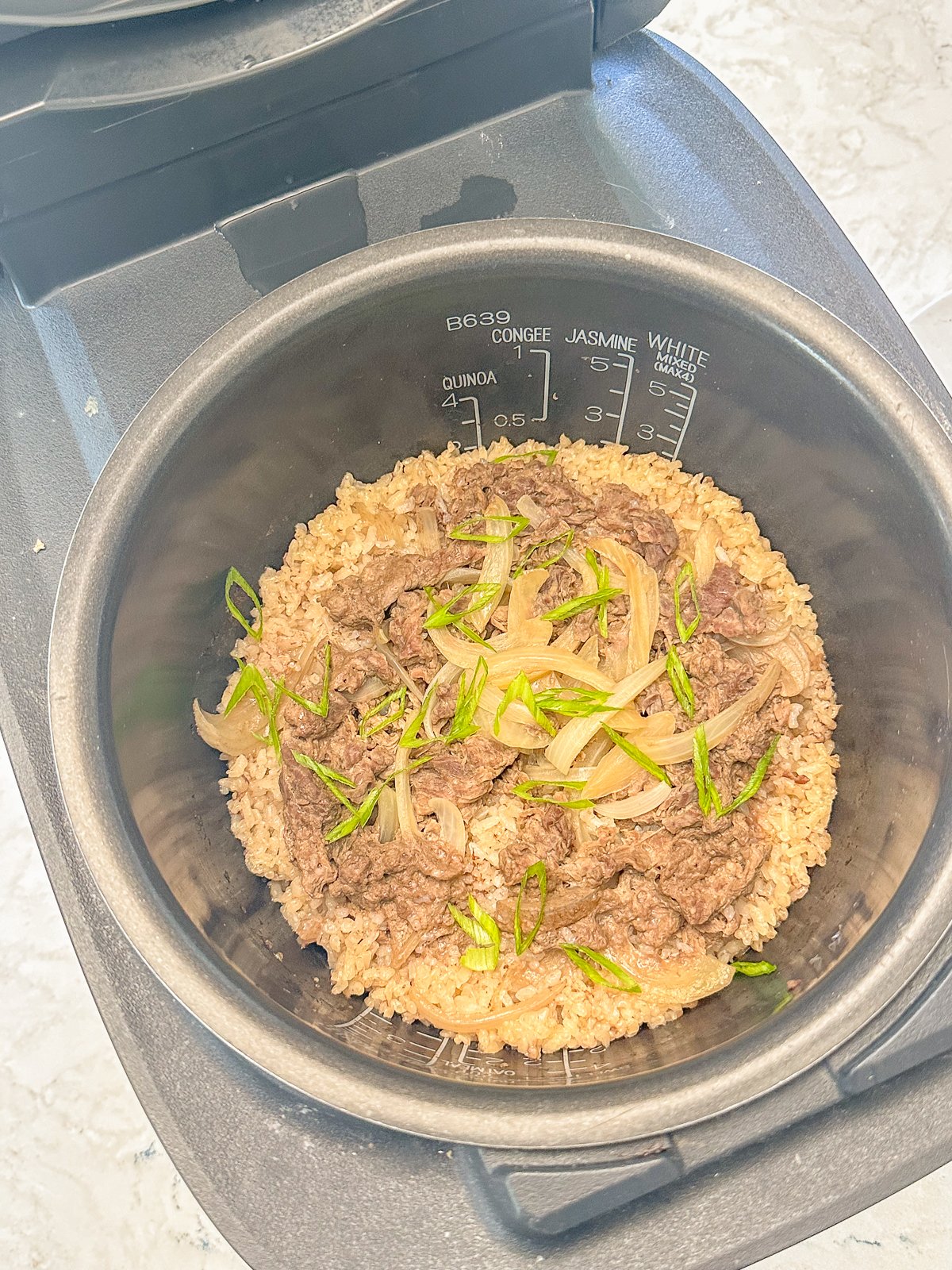 Rice cooker filled with cooked rice, tender beef slices, and white onions, topped with fresh green onions.