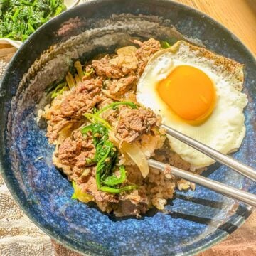 Beef and rice bowl with onions, served with a fried egg, garnished green onions, and steamed vegetables on the side.