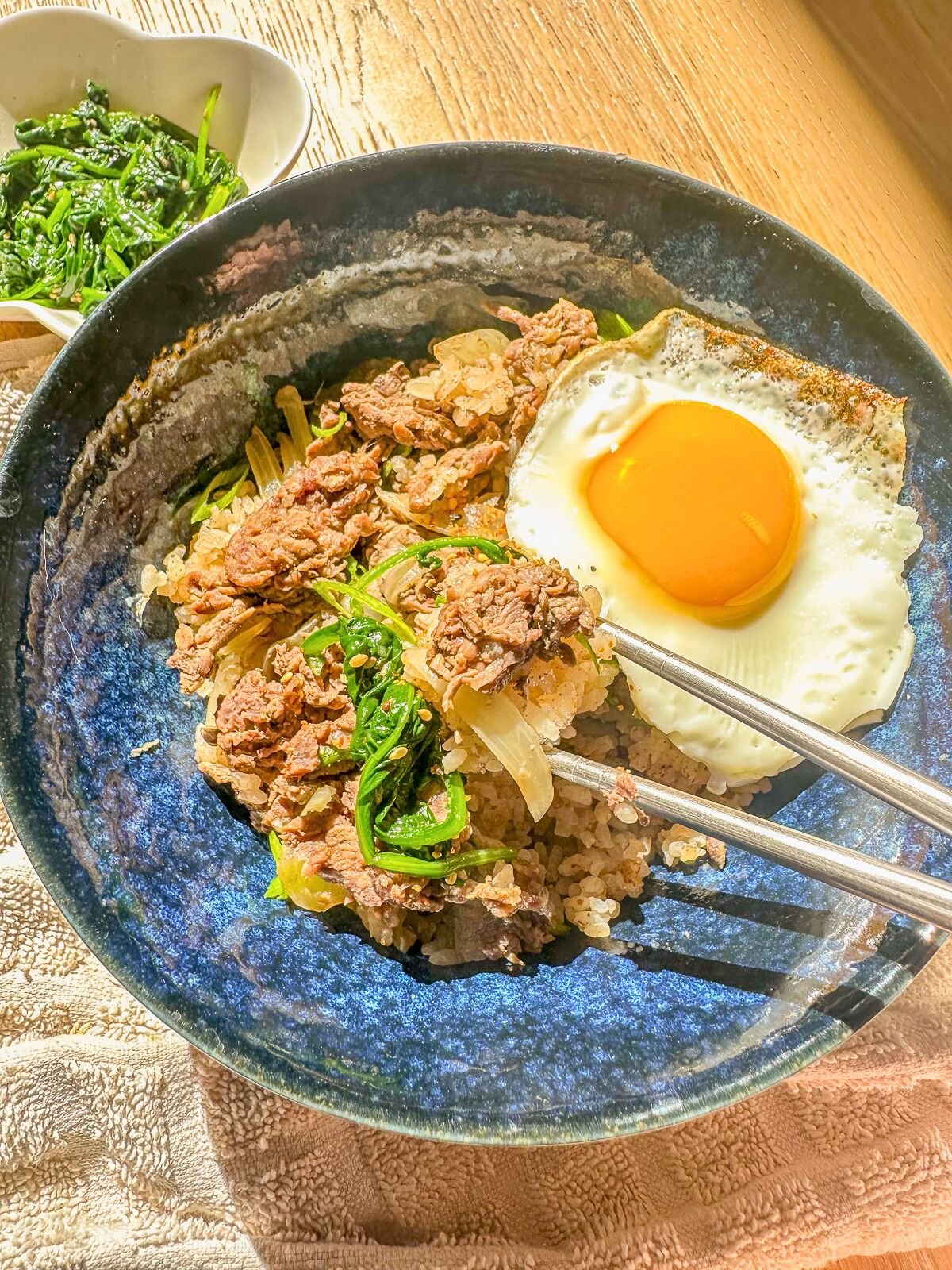 Beef and rice bowl with onions, served with a fried egg, garnished green onions, and steamed vegetables on the side.