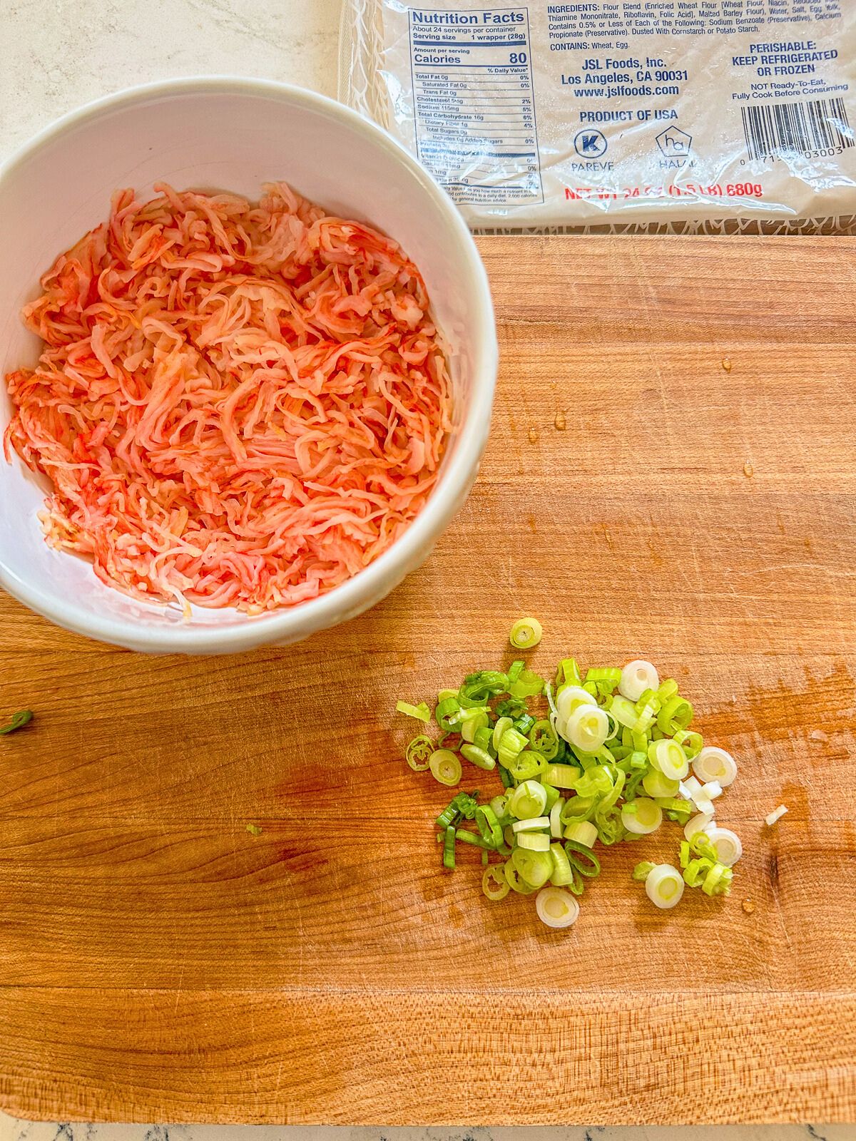 Wooden chopping board with a bowl of imitation crab and fresh chopped green onions on the side.