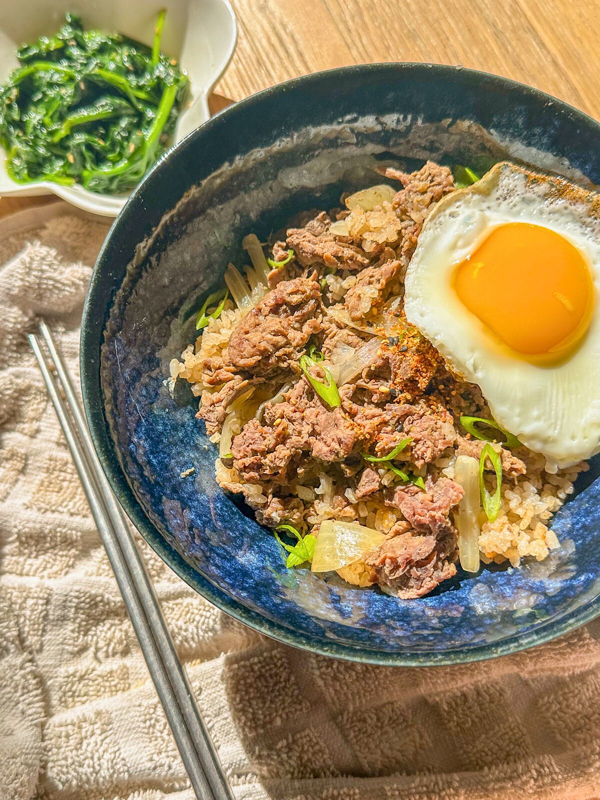 Beef and rice bowl served with a fried egg, garnished with green onions, and steamed vegetables on the side.