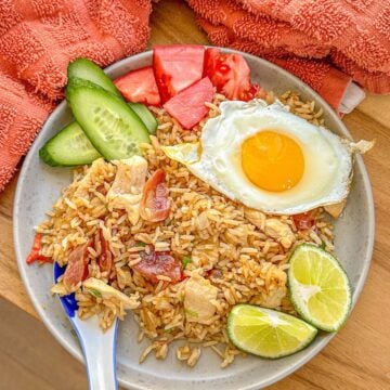 A plate of fried rice with fried egg, slices of cucumbers, tomatoes and lime on the side.