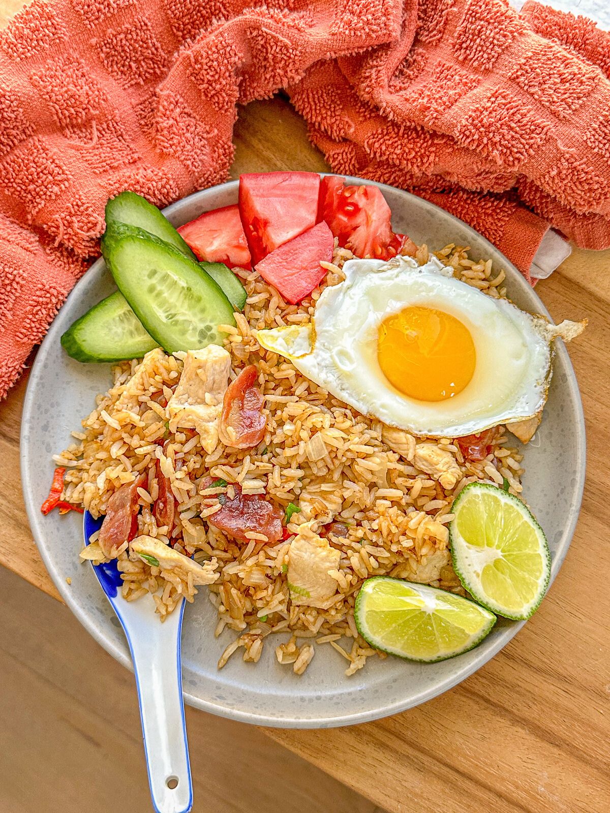 A plate of fried rice loaded with chicken and Chinese sausage, served with a fried egg, cucumber, tomato, and lime on the side.