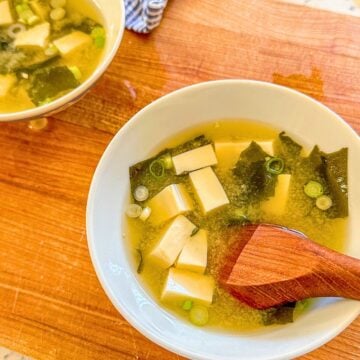 Miso soup in a bowl on a wooden board with tofu, dried seaweed, green onions, a wooden spoon, and another bowl of soup beside it.