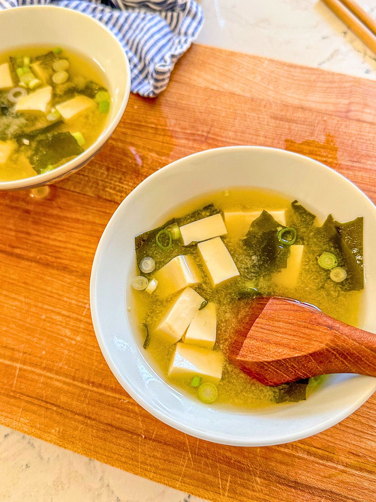 Miso soup in a bowl on a wooden board with tofu, dried seaweed, green onions, a wooden spoon, and another bowl of soup beside it.