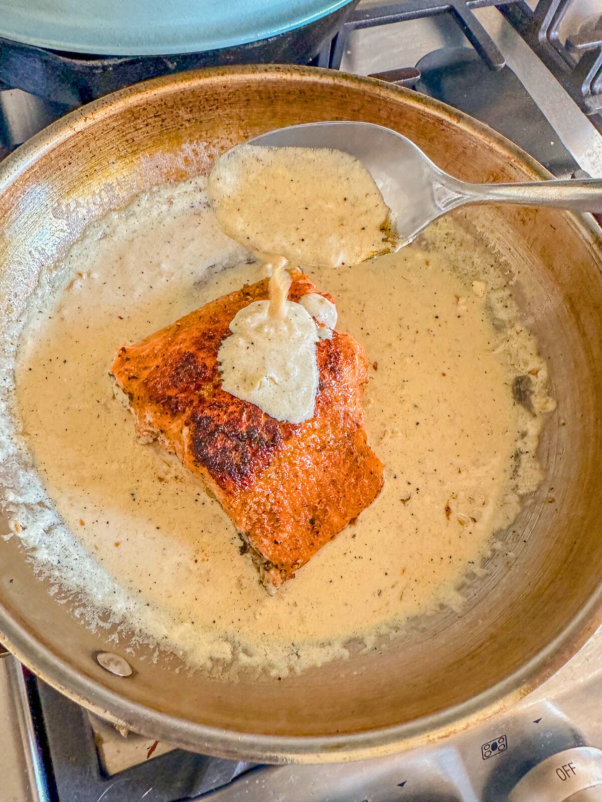 Cream sauce being poured over salmon simmering in a pan of sauce.