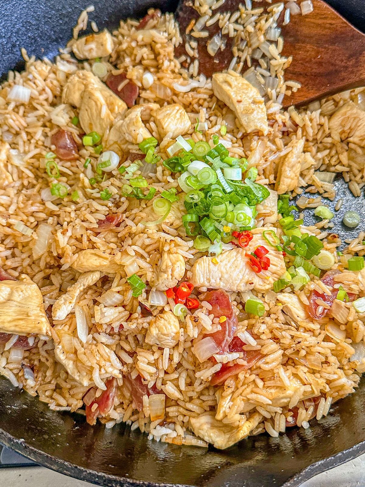 Fried rice in a pan with onions, garlic, Chinese sausage, and chicken, coated in sweet soy sauce and topped with green onion.