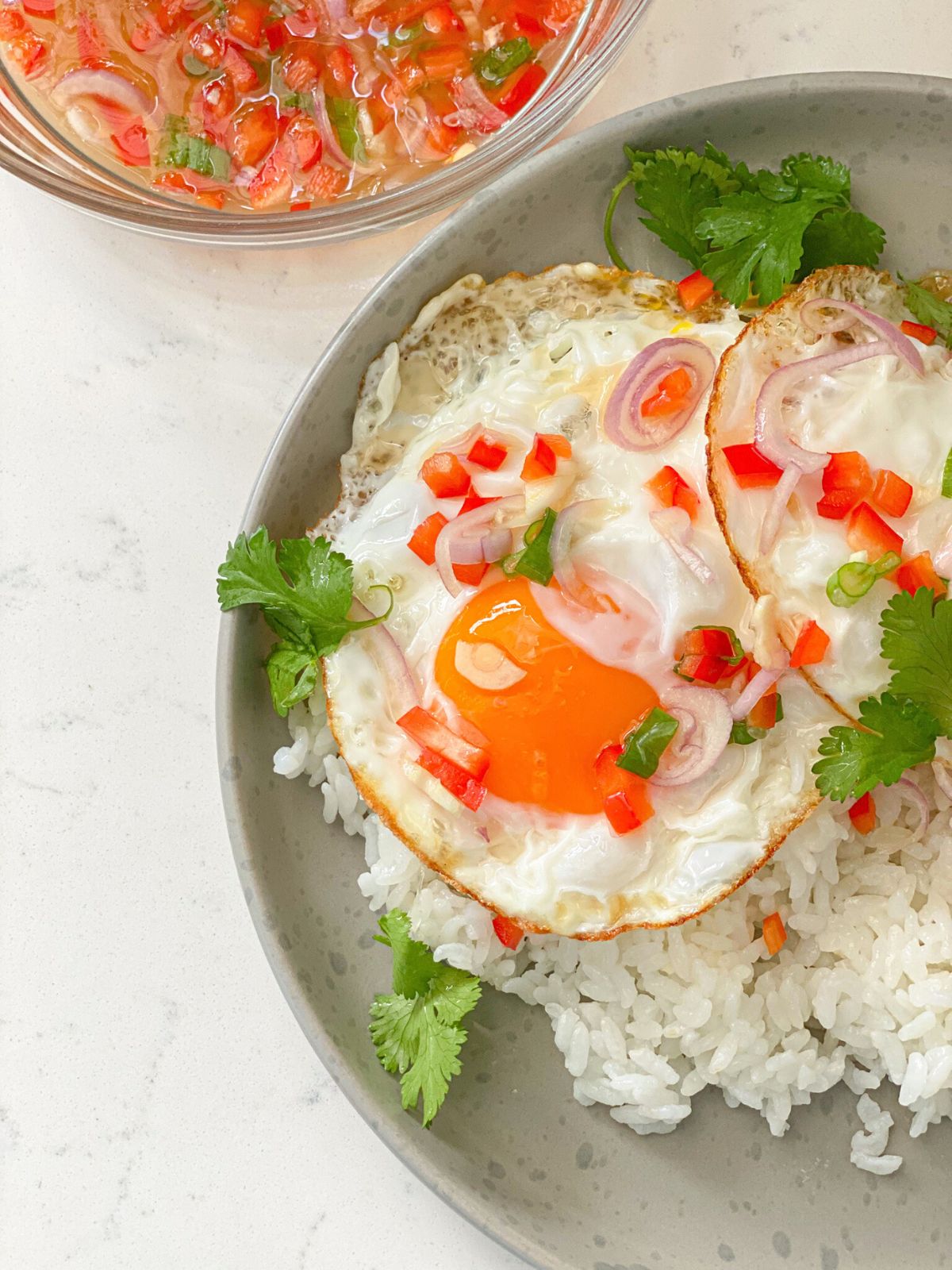 Sunny-side-up eggs over white rice, garnished with shallot, scallion, red bell pepper and cilantro served with a lime fish sauce on the side.