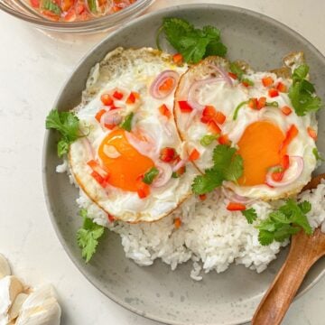 Sunny-side-up eggs over white rice, garnished with shallot, scallion, red bell pepper and cilantro served with a lime fish sauce on the side.
