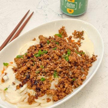 A plate of udon noodles with ground pork in creamy sauce, garnished with green onions, placed beside a jar of chili oil and chopsticks.