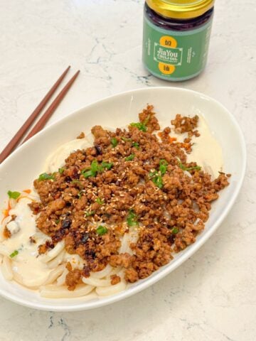 A plate of udon noodles with ground pork in creamy sauce, garnished with green onions, placed beside a jar of chili oil and chopsticks.