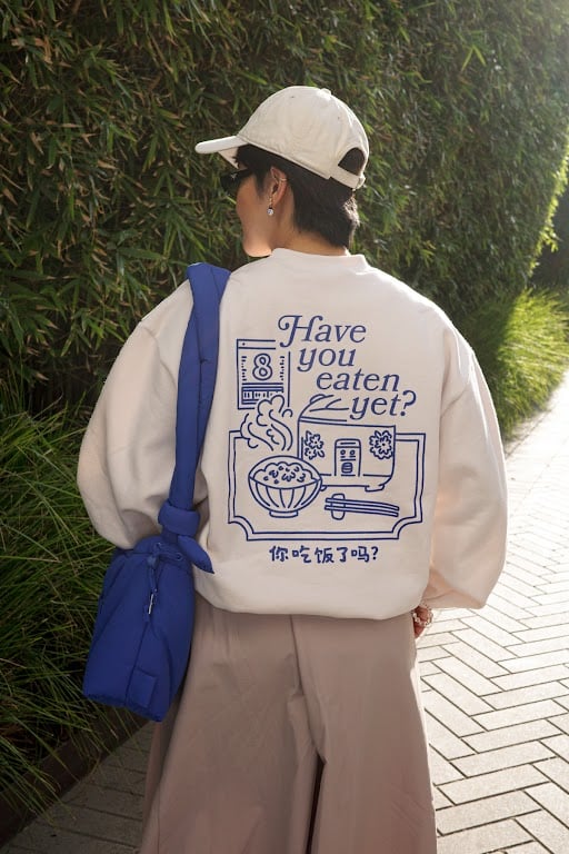 Person facing away, showing her back, wearing a cream crewneck sweater, cream cap and a blue shoulder bag.