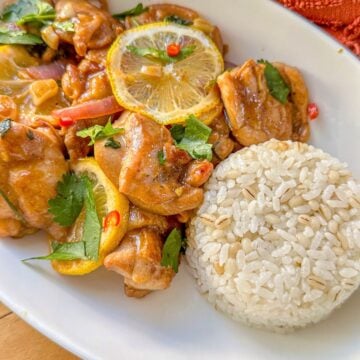 A plate of cooked chicken topped with lemon slices, chopped onions, cilantro, and chilies served with white rice.