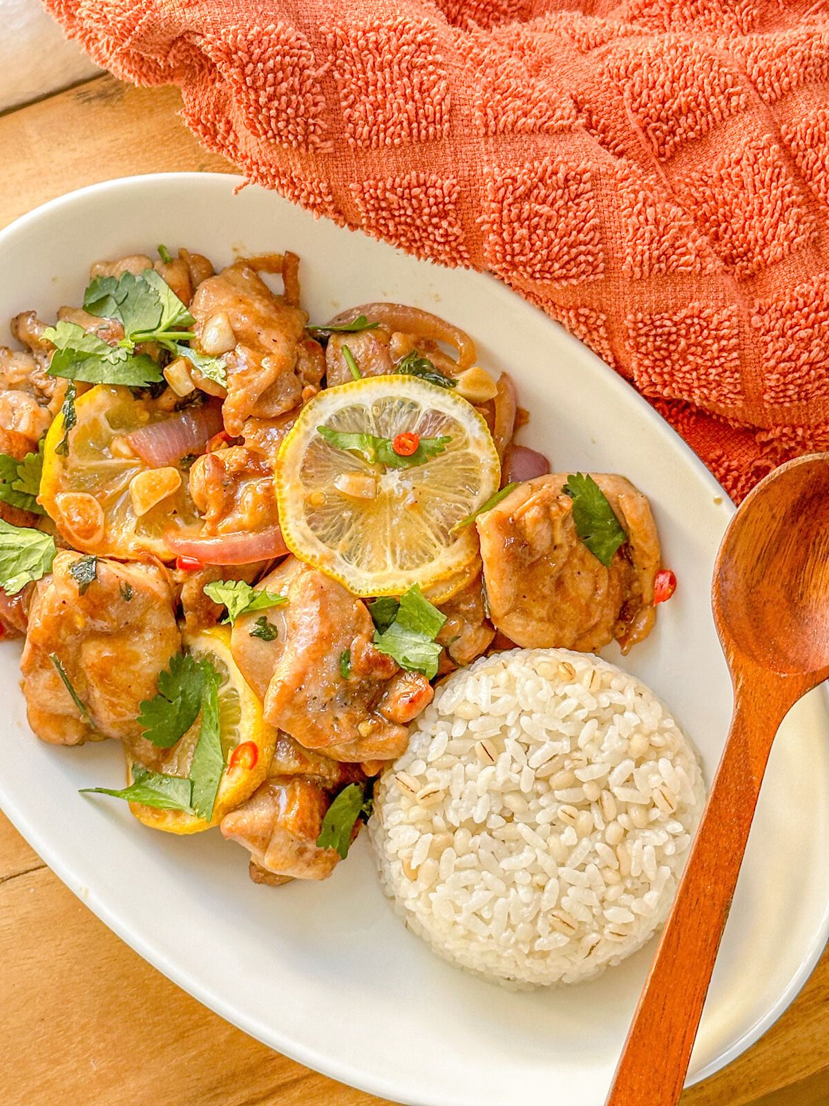 Cooked chicken topped with lemon slices, chopped onions, cilantro, chilies and served with white rice, and a wooden spoon on the side.