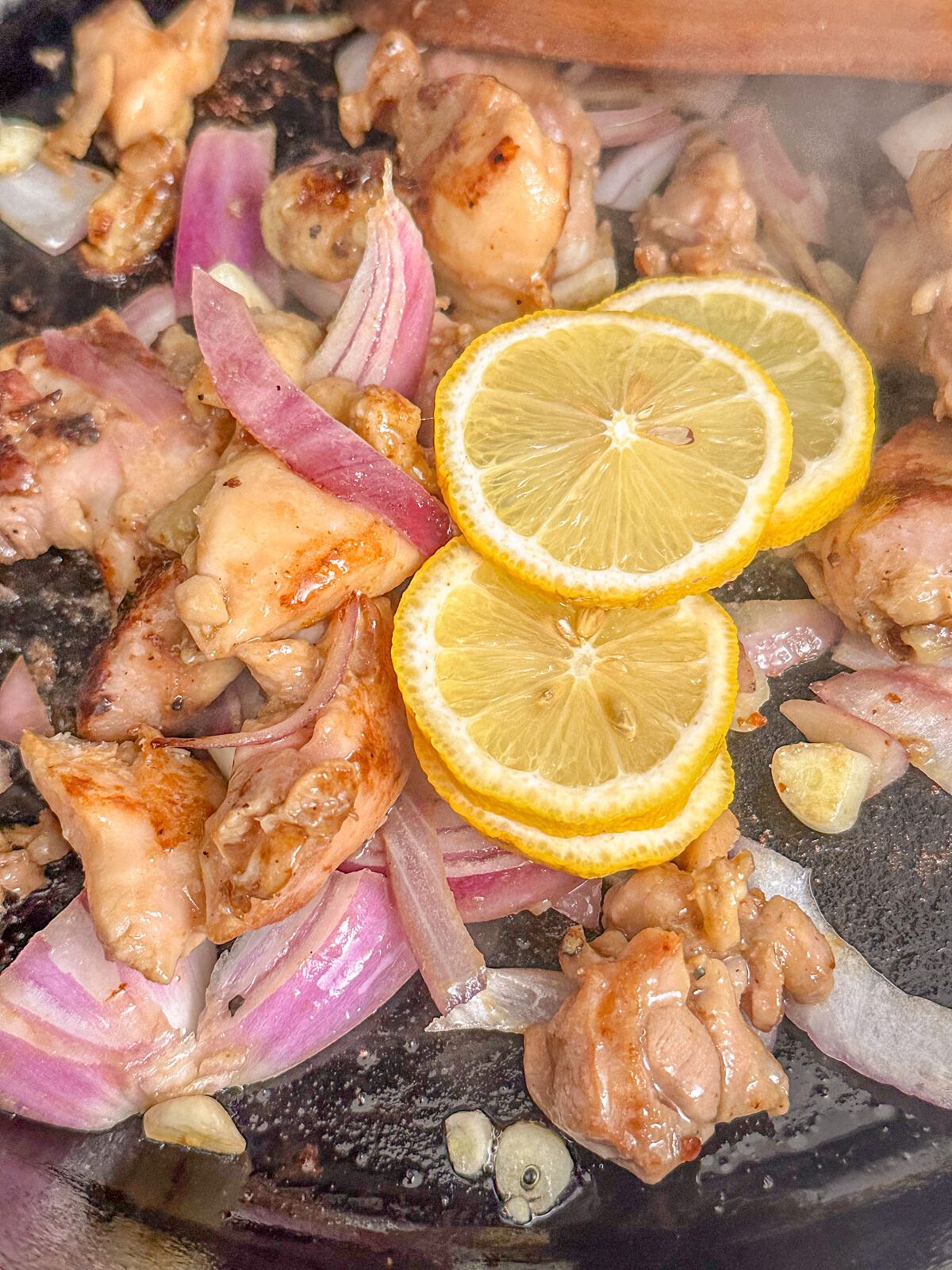 Chicken cooking in a pan with chopped onions, garlic, and lemon slices.