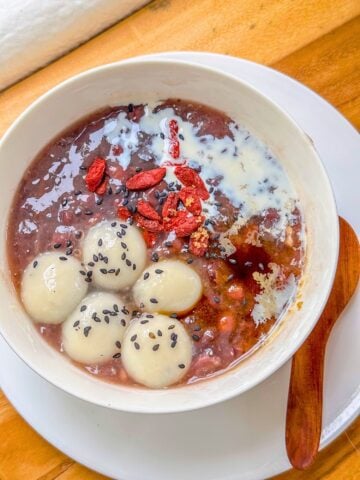 A bowl of red bean soup topped with glutinous rice balls, goji berries, sesame seeds and a light drizzle of milk.