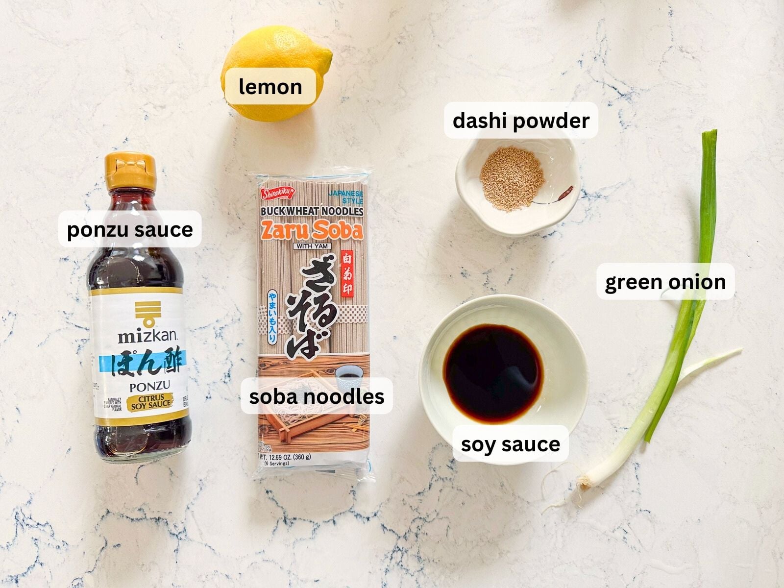 Ingredients for hot soba noodles laid out on a table countertop including lemon, dashi powder, green onion, soba noodles pack and ponzu sauce.