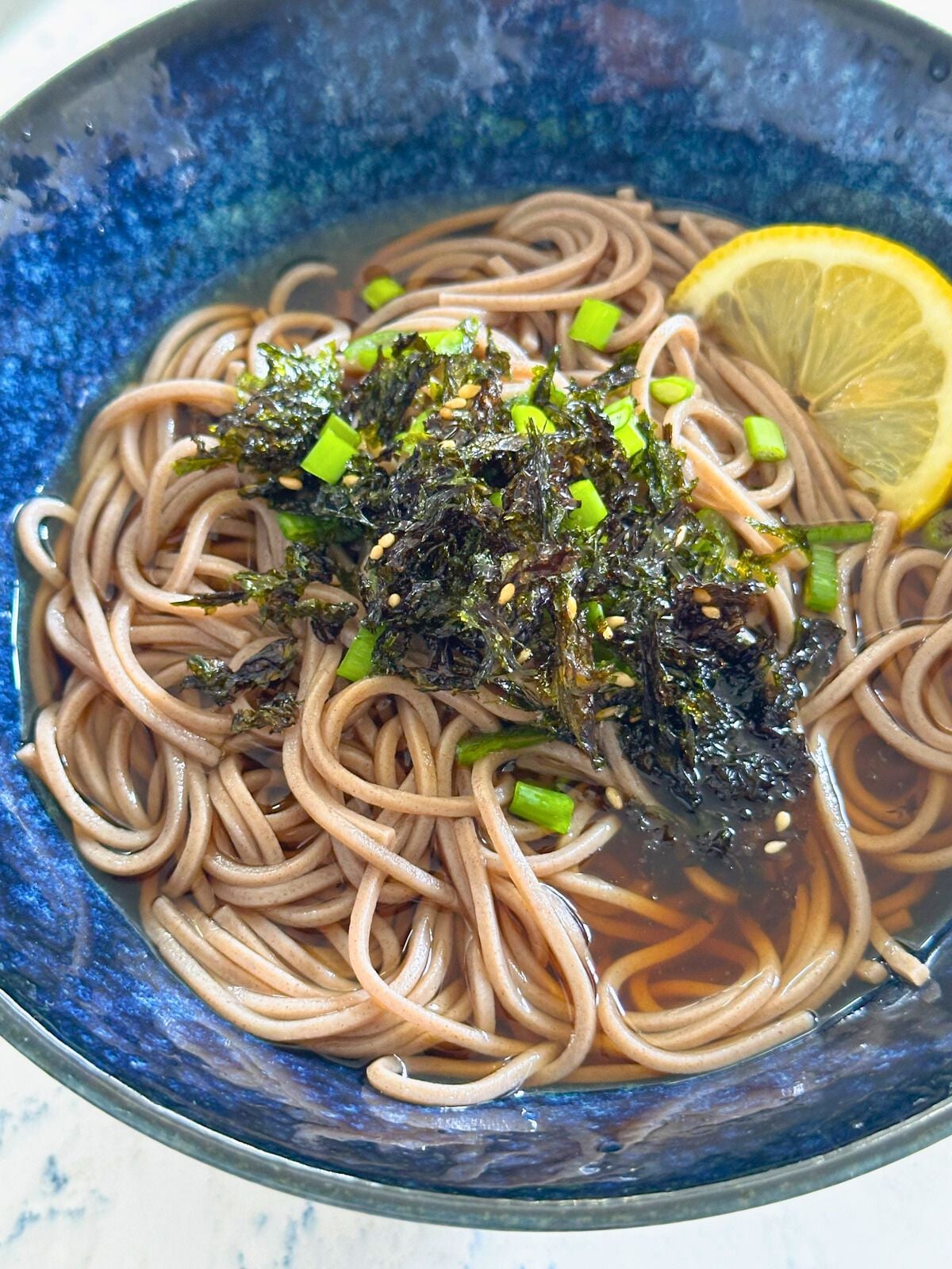 A bowl of cooked soba noodles topped with sliced lemon, green onions, nori and sesame seeds.