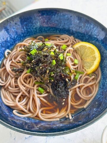 A bowl of cooked soba noodles topped with sliced lemon, green onions, nori and sesame seeds.