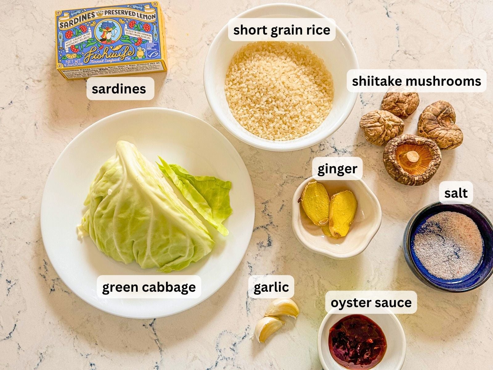 Ingredients laid out on a marble table countertop including short grain rice, sardines, green cabbage, ginger, mushrooms, garlic, salt and oyster sauce.