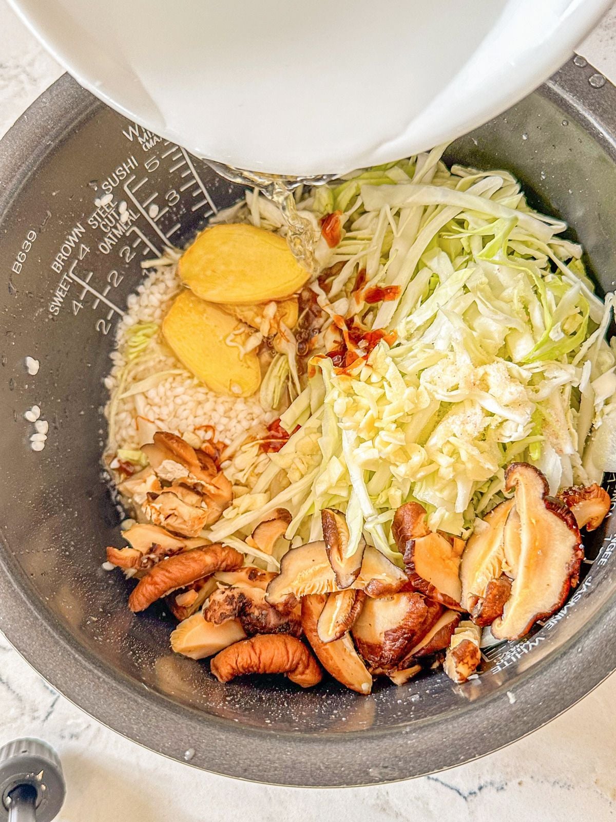 A pot of uncooked rice with water, sliced ginger, shiitake mushrooms and cabbage.