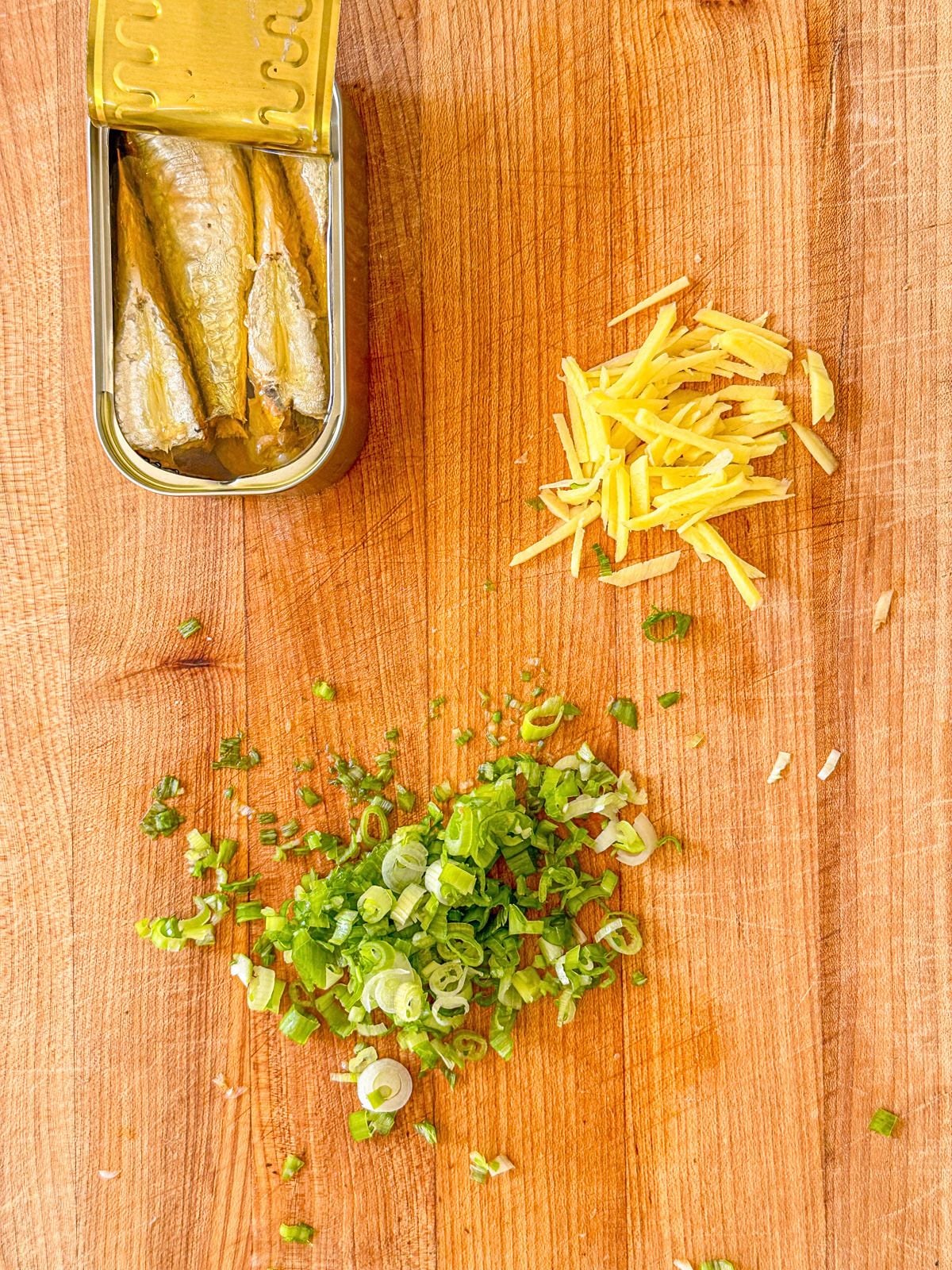 Sliced green onions and ginger on a chopping board with an open can of sardines.