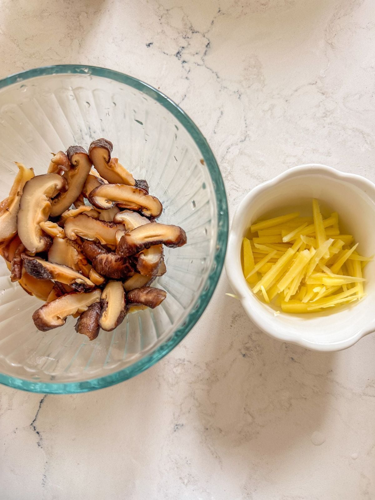 Sliced mushrooms in one bowl beside peeled ginger cut into thin strips in another bowl.
